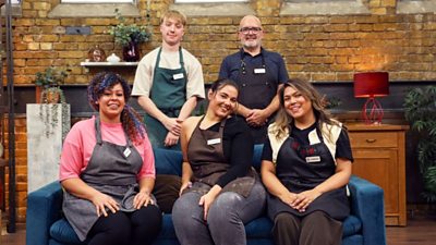 A group shot of the MasterChef contestants smiling and wearing aprons. They are on or around a blue sofa, and in front of an exposed brick wall.