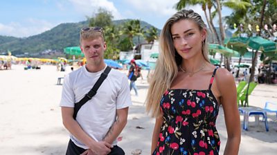 Zara and Loukas stood on a sunny beach looking at the camera. There are some parasols and sunbeds behind them.