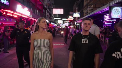 Zara and Mac walking down a street at night that is filled with nightclubs, bars and neon lights.