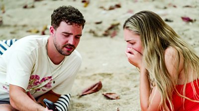 Zara and Hugo lying on a sandy beach. He is showing her something on his mobile phone and she has her hand over her mouth in an expression of shock