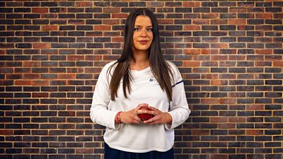 A young woman in a white long sleeve top stands against a brick wall holding a cricket ball