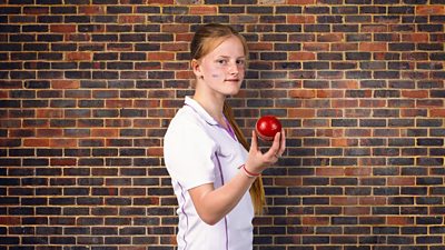 A young woman in a short sleeved white shirt stands holding a cricket ball