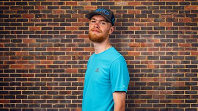 A young man stands against the backdrop of a wall wearing a light blue t-shirt and dark blue baseball cap