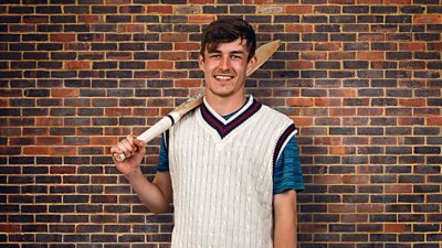 A young man wears a white sleeveless cricket jumper over a blue t-shirt and holds a cricket bat, slung over his shoulder