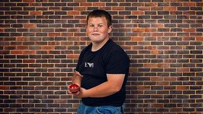 A young man in a black t-shirt and jeans stands by a wall holding a cricket ball