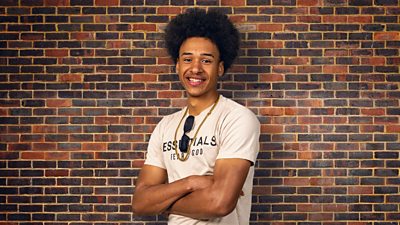 A young man in a light t-shirt stands by a wall with his arms folded, smiling to camera