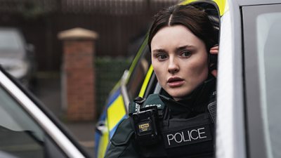 A woman in police uniform leans out the door of a police car looking concerned