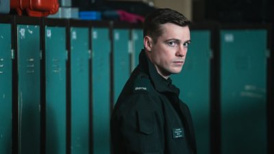 A man in police uniform stands in a locker room looking wary of something off screen