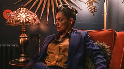 A woman sits in a decadent armchair looking suspicious of something off screen