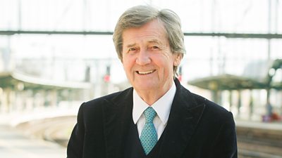 Headshot of Melvyn Bragg. He wears a suit and smiles to the camera while standing outdoors in a sunny location.