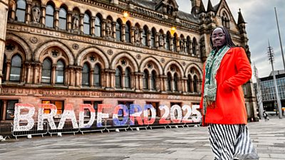 A woman in an orange jacket and black and white skirt stands in Bradford city centre beside a red white and blue sign for Bradford 2025
