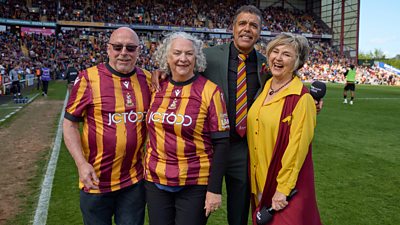 Bantam of the Opera choir members with Chris Kamara and Lesley Garrett, standing on a football pitch