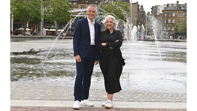 A man in a dark suit and a woman in a black shirt and skirt stand together smiling in front of a fountain