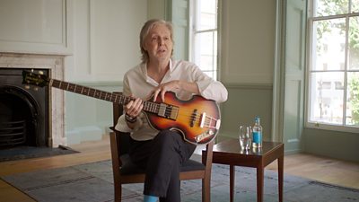 Paul McCartney sits on a chair and rests a guitar on his knee. Leaning forward, he is mid-conversation with someone off-camera. He is sat in a large living room.