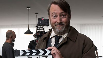 David Mitchell on the set of Ludwig. Holding a clapperboard, he smiles to the camera. Camera crew are visible in the background.