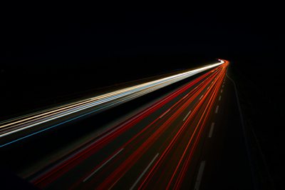 An abstract image of some lights (presumably from a car or traffic) blurred across the frame as they moved too fast for the shutter, creating multiple thin and brightly lit red and white lines on a very dark black background.