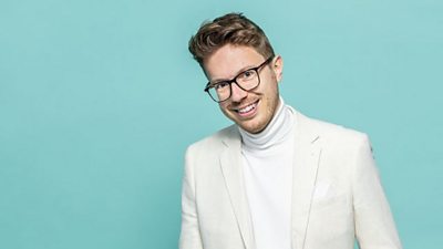 James B Partridge with school hymn book against a light blue background. He is wearing a very pale blazer jacket with a white turtle collared top underneath it.