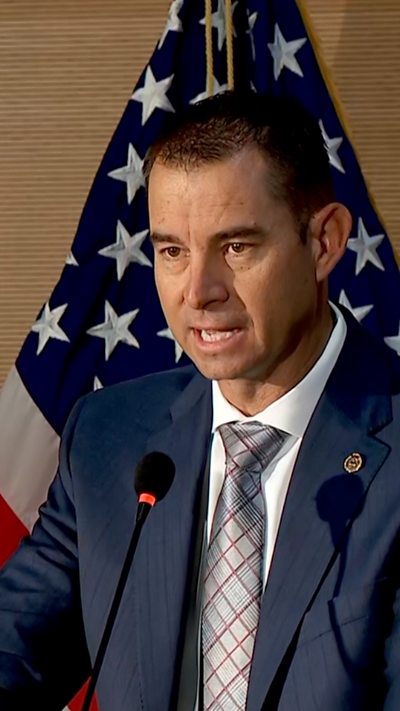 A man wearing a blue suit, white shirt and silver striped tie speaks into a microphone in front of an American flag.