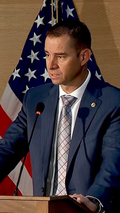 A man wearing a blue suit stands at a podium in front of an American flag.