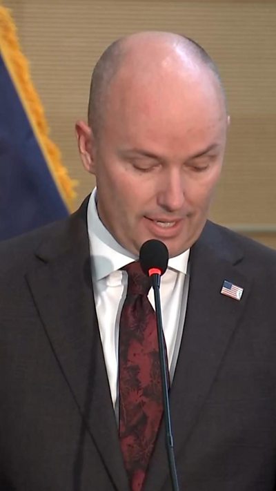 A bald man speaks into a microphone wearing a blue suit, white shirt and red tie.
