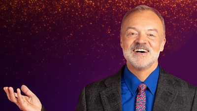 A man in a grey suit jacket, blue shirt and floral tie looks to camera smiling and laughing against a purple backdrop, flecked with gold