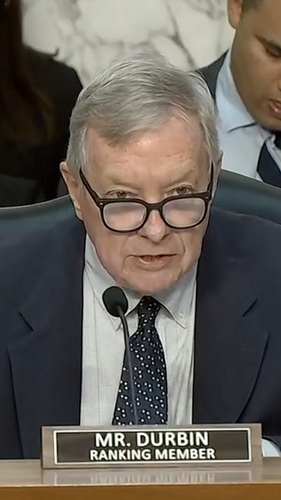 Dick Durbin is shown talking into a microphone behind a large dias in a US Senate hearing room.
