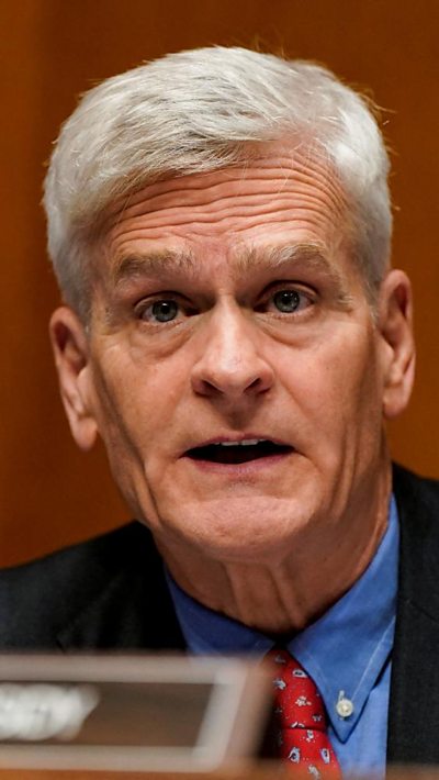 A man wears a blue suit, blue shirt and red polka dot tie while speaking toward a witness while sitting behind a dias.