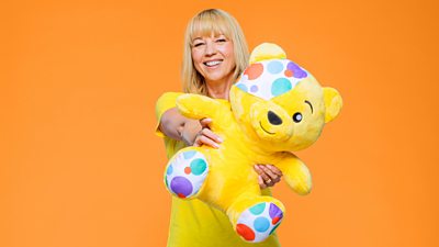 Sara Cox wearing a Children In Need yellow t-shirt and holding a toy Pudsey bear out towards the camera. She is smiling against an orange background.