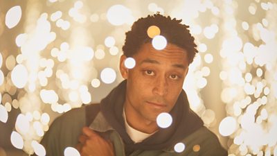 Ben Coyle-Larner looking at the camera surrounded by warm yellow lights. His hand is holding a bag on his right shoulder.