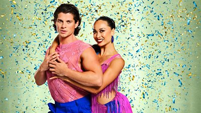 Lewis wears a sleeveless pink top and blue trousers while Katya wears a blue and purple cutout dress with purple fringe trim