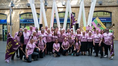 Approximately 30 people in a group shot wearing pink shirts in the concourse of a train station. A few of them are holding large Bantom of the Opera flags.