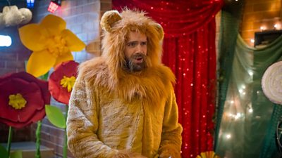 A man in a lion suit stood in front of some large papier mache flowers, a sparkly red curtain and some fairy lights