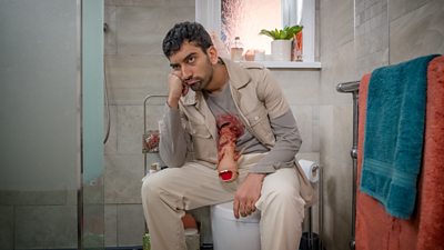 A man sat on a toilet his face resting on his fist. He has a fake wound going into his chest.