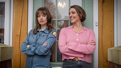 Two women stood leaning against a front door pained with glass and a wooden frame.