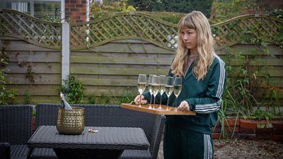 A young girl in a tracksuit carrying a tray of champagne glasses across a garden towards some wicker furniture.