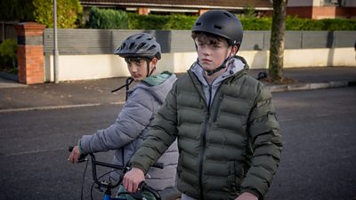 Two young boys on bikes, wearing safety helmets, on a road.