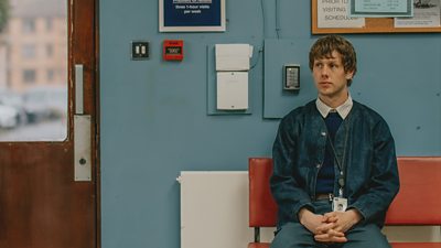A man sits in a waiting room on a bench. He wears a lanyard over a denim jacket, jumper and jeans. 