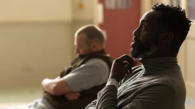 Close-up of a man sat on a wooden chair in a community hall. 