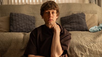 Close-up of a man sat on the floor, leaning against a sofa. He stares directly at the camera. 
