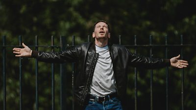 A man stands outdoors with his arms outstretched and his head facing up towards the sky. 