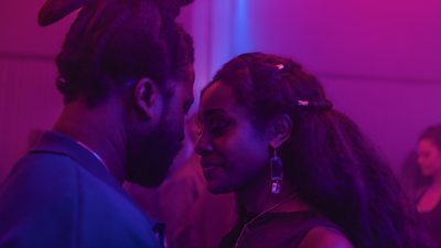 A young man and a woman stood face to face with their eyes closed in a dark room with a pink hue.