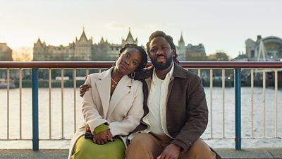 A man and woman sat on a bench at the waterfront of the Rover Thames.  His arms is around her shoulders.  The water and sky is grey.