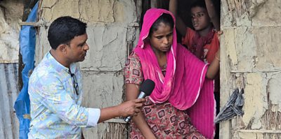 A male journalist in Bangladesh, interviewing a local woman for a newspaper article - she is wearing a pink head scarf, he is wearing a light blue shirt with a pattern and earring a microphone. They are at her home, one of her children stands behind her in the doorway