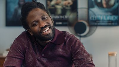 Adjani Salmon in character as Kwabena sat at a table with film posters on the wall behind him. He is smiling.