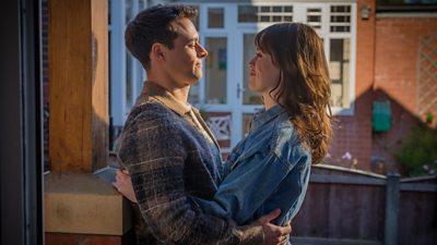 A young man and woman embracing with a red brick house behind them. They are smiling.