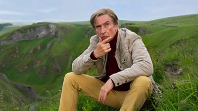 A man in a beige jacket and mustard yellow trousers sits in grass at the top of a very tall hill looking contemplative