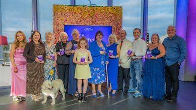 A group shot of twelve people and one dog. Some of which are holding purple trophies. They are in front of a Make a Difference branded screen.