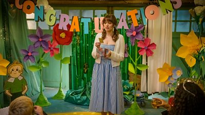 Aimee Lou Wood stood under a Congratulations banner made out of paper.  She is wearing a blue and white gingham dress. There are large papier mache flowers around her
