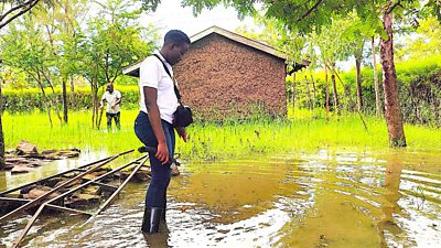 a journalist standing in flood water outside a small house in Kenya