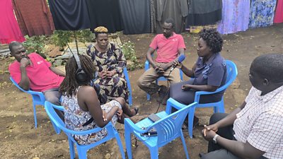 Journalist is sitting with five people from a rural community in Kenya, they are sitting on blue plastic chairs and washing is hanging on the line behind them.